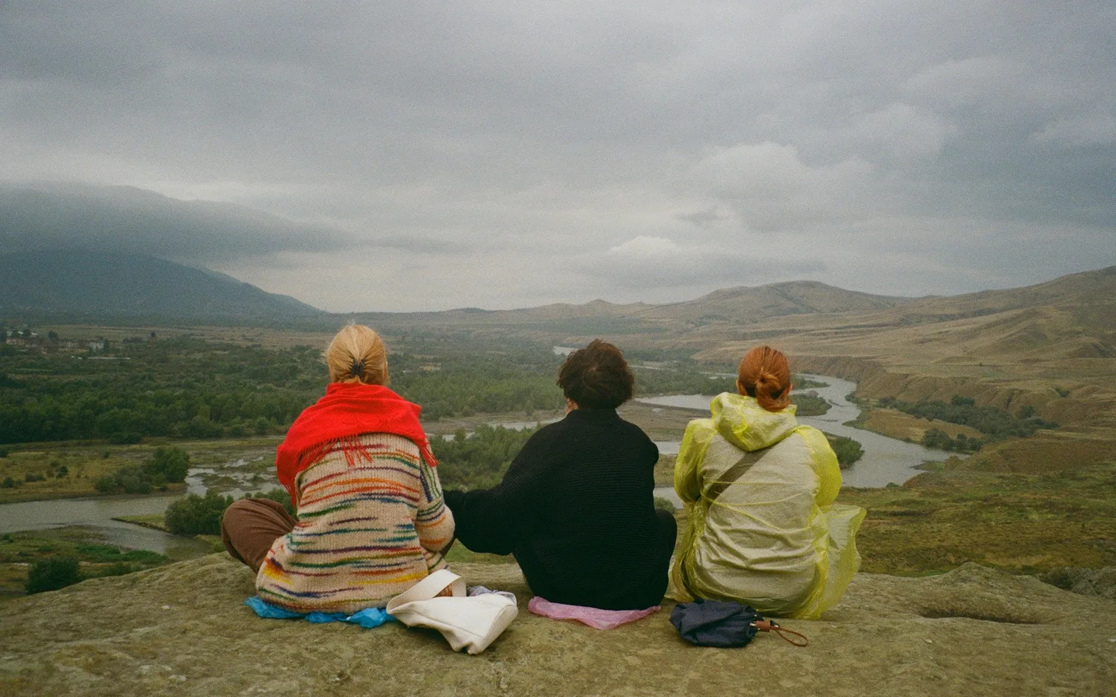 Four travellers with backpacks walking a mountain trail together, seen from behind, heading toward snow-covered peaks.