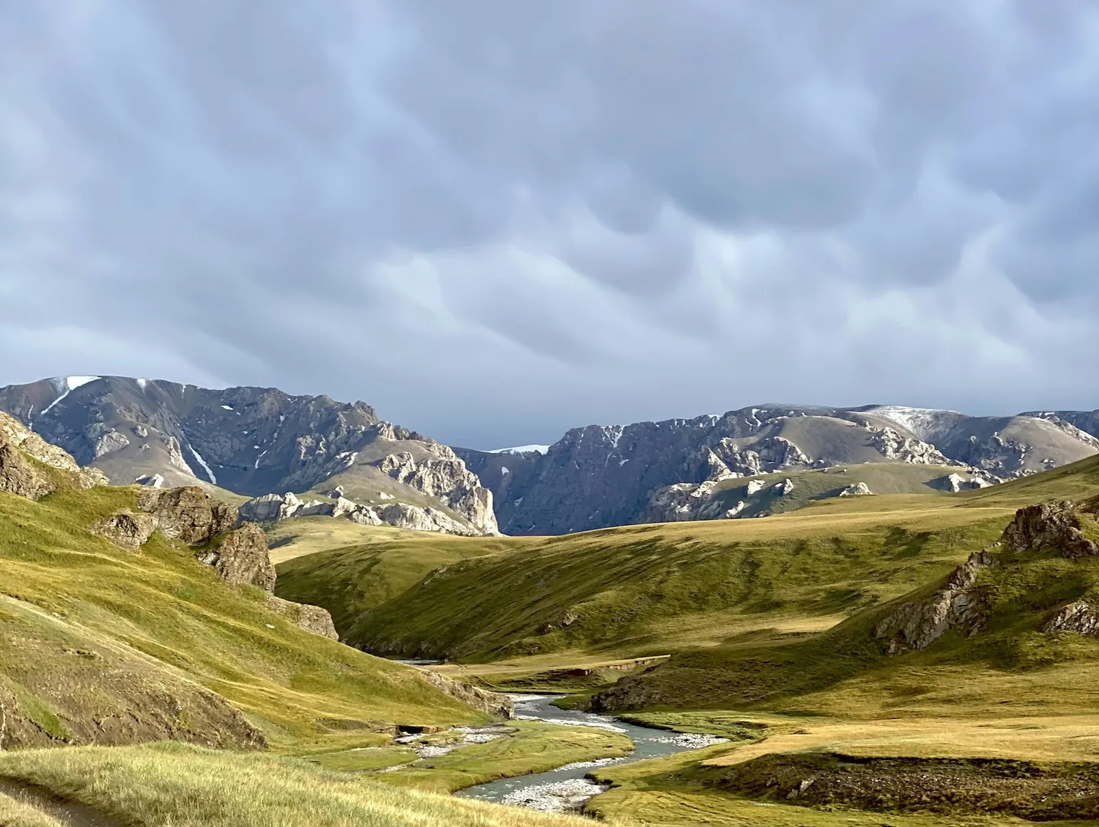 A green mountain valley in Kyrgyzstan, a river winding below snow-dusted ridges under heavy cloud.