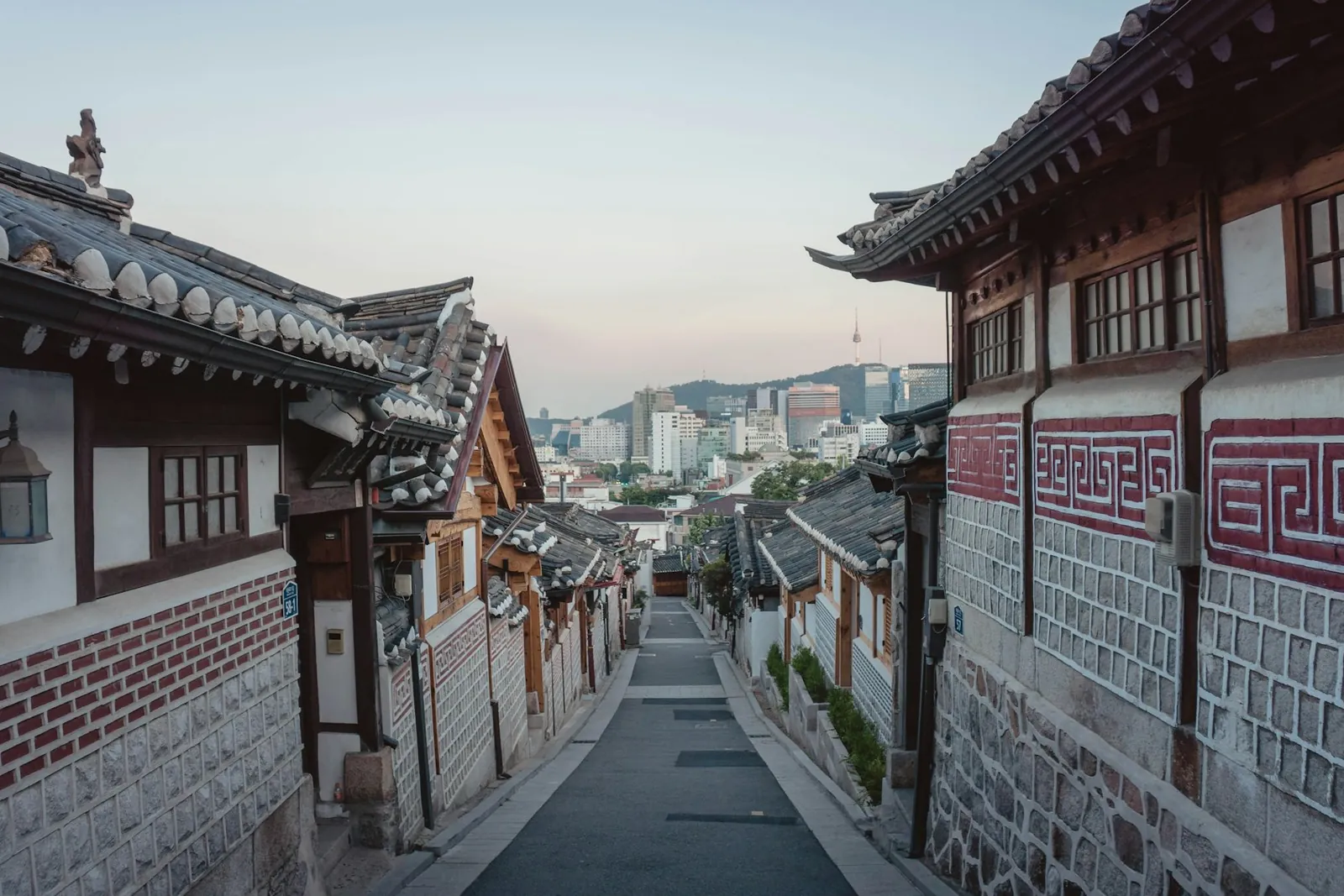 A lane of traditional tiled hanok roofs in Bukchon, with the towers of modern Seoul rising behind.