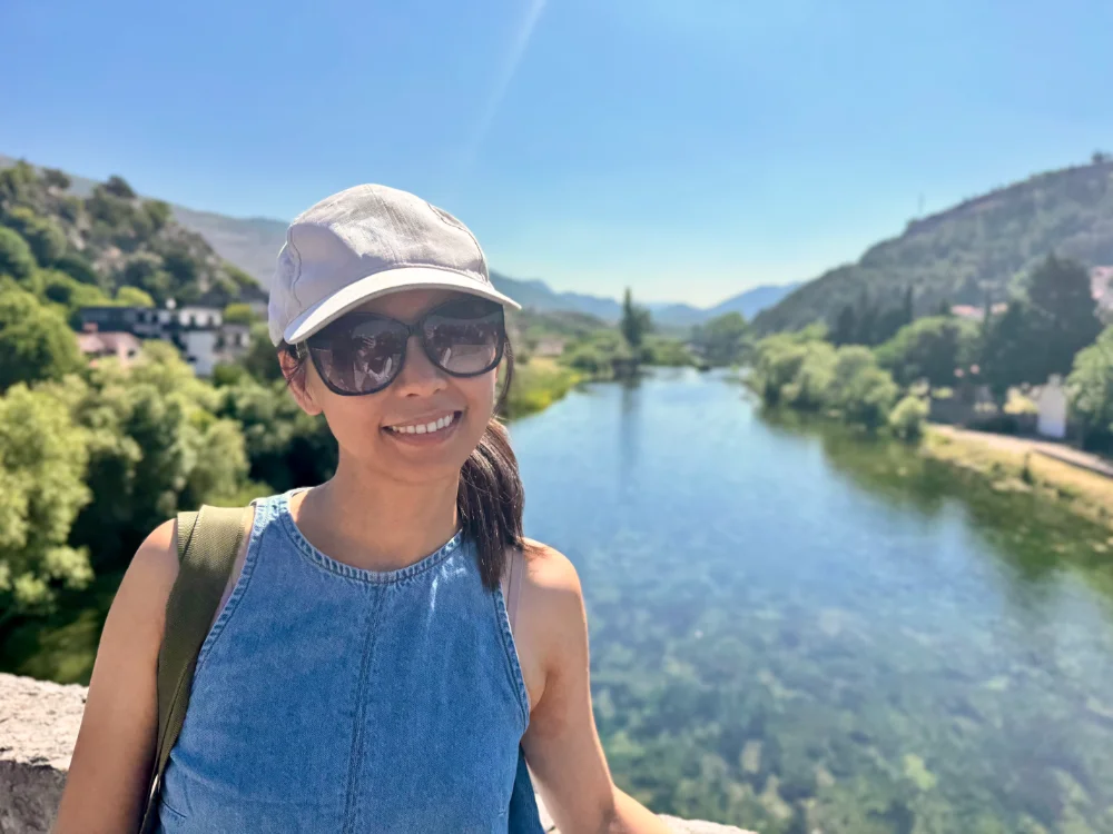 Ting-Yi Shih, founder of Shih Travels, smiling in a cap and sunglasses on a bridge above a turquoise river valley.